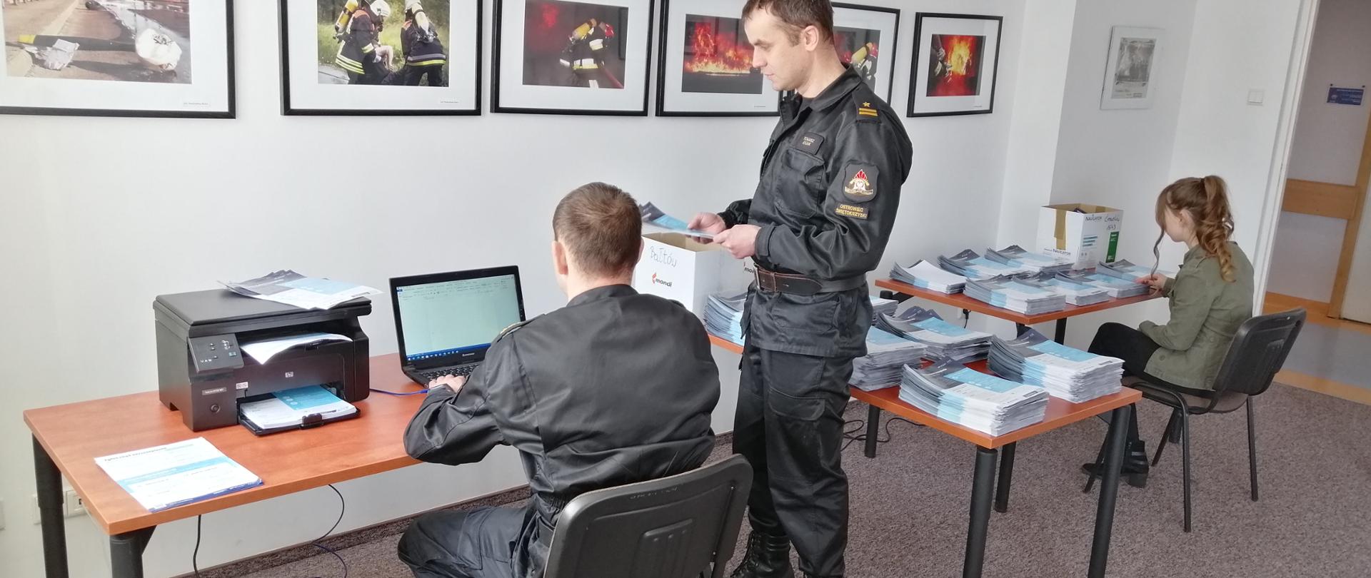 Na zdjęciu widać pokój biurowy. Na pierwszym planie strażak drukuje ulotki. Na drugim planie kobieta układa wydrukowane ulotki na stoliku. Pracę nadzoruje Zastępca Komendanta Powiatowego PSP w Ostrowcu Św. 