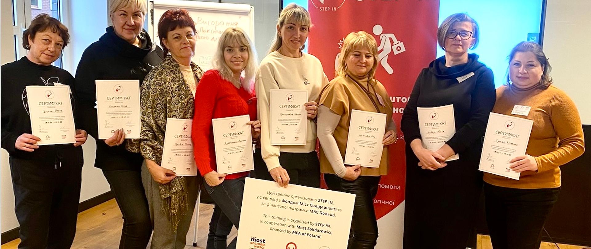 8 kobiet pozuje do zdjęcia z certyfikatami i planszą z informacją, że projekt został zrealizowany przez Step In, Most Solidarności z finansowaniem MFA of Poland