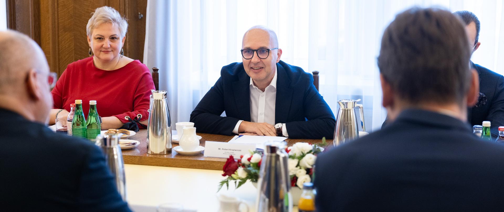 Wiceminister Robert Kropiwnicki siedzi za stołem obok kobiety i mężczyzny. Na przeciwko siedzi dwóch mężczyzn, widać tylko ich plecy. W tle biała zasłona. Na stole filiżanki, dzbanki i kwiaty. 