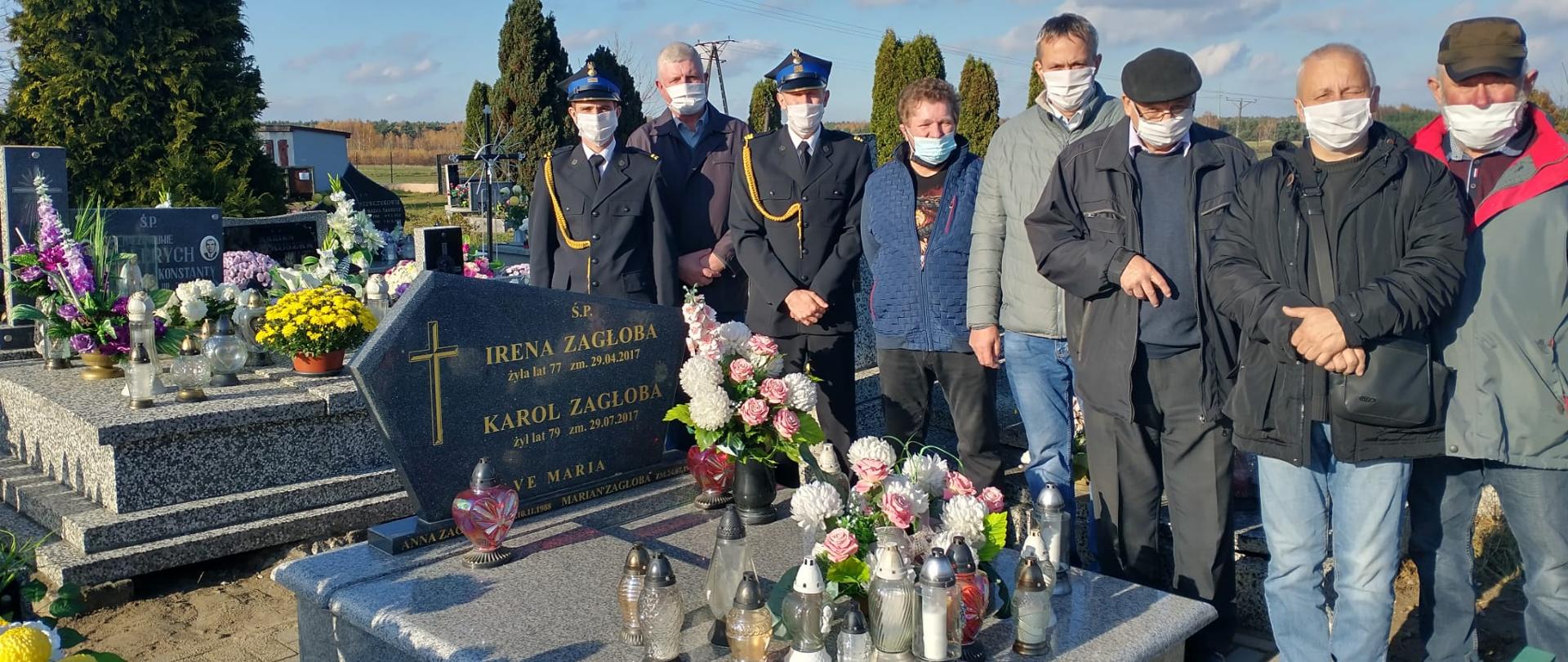 Dwaj strażacy ubrani w mundurach stoją wraz z sześcioma mężczyznami na cmentarzu przy pomniku na którym ustawione są znicze i kwiaty. Wszyscy mają założone maseczki Na płycie pomnika widać napis śp Irena Zagłoba żyła lat 77 Karol Zagłoba żył lat 79