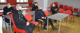 Świetlica Komendy Powiatowej PSP w Rawiczu. Przy stolikach na krzesłach siedzi 5 mężczyzn, w tym 4 umundurowanych strażaków. Uczestniczą w szkoleniu z obsługi symulatora zagrożeń. W tle okna oraz flagi.