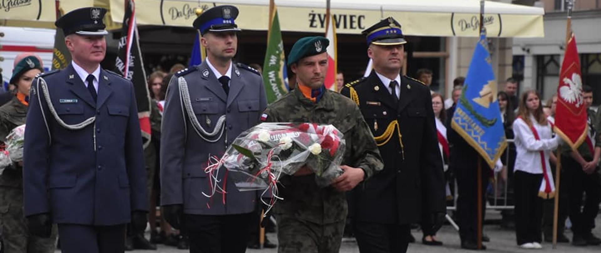 Zdjęcie przedstawia 4 mężczyzn, którzy maszerują obok siebie. Pierwszy w mundurze moro niesie wiązankę kwiatów. Pozostali trzej w różnego koloru mundurach galowych maszerują za nim. W tle widać zgromadzenie ludzi, sztandary, fasady budynków i banery reklamowe.