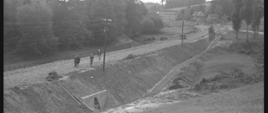 Czarno-białe zdjęcie, na którym widoczni są robotnicy stojący na uklepanej drodze. Wzdłuż drogi ciągnie się skarpa z jednym przepustem.