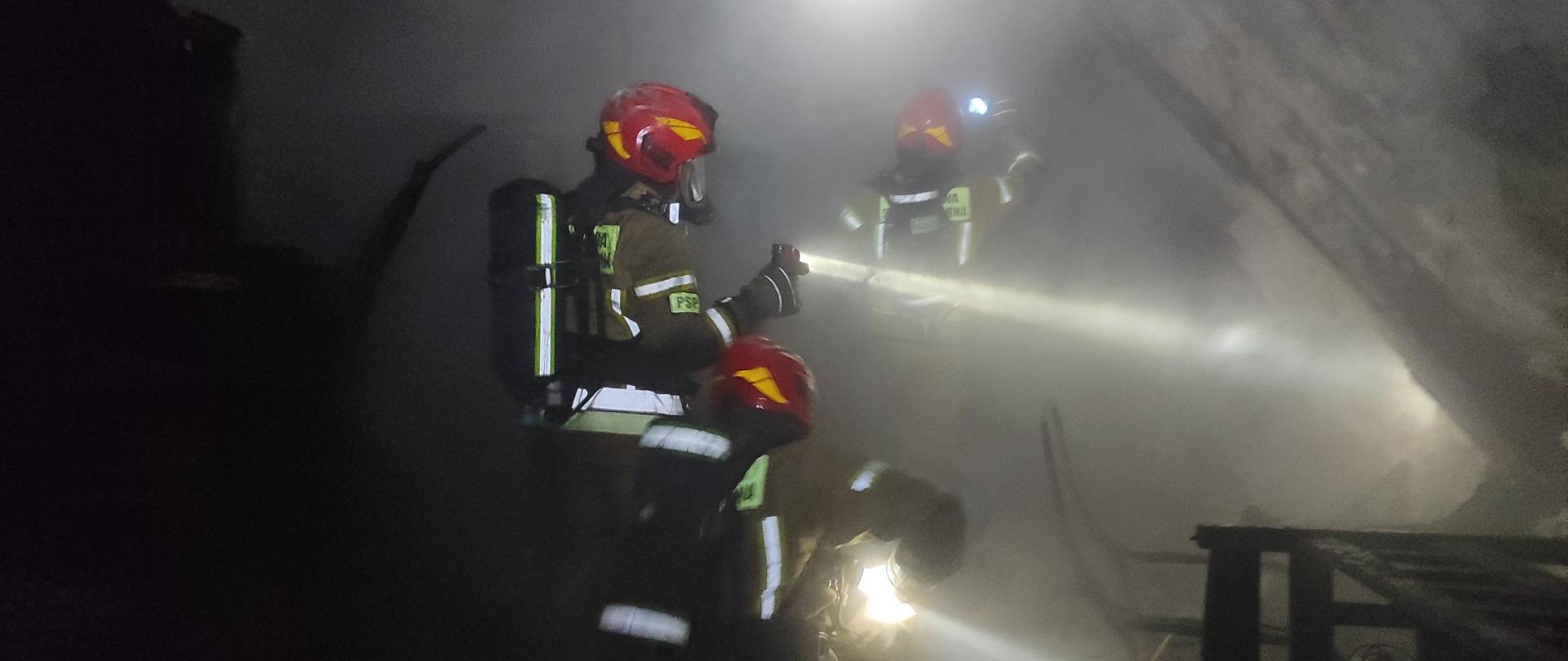 Zdjęcie przedstawia strażaków w trakcie działań gaśniczych w silnie zadymionym pomieszczeniu. Trzech ratowników ubranych w specjalistyczne ubrania ochronne i hełmy z oznaczeniem PSP (Państwowa Straż Pożarna) prowadzi działania z użyciem linii gaśniczej. Pomieszczenie jest zaciemnione i wypełnione gęstym dymem, co wskazuje na zaawansowany pożar w warunkach zadymienia. Ratownicy poruszają się ostrożnie i metodycznie, prawdopodobnie przeszukując pomieszczenie lub dogaszając zarzewia ognia. Zdjęcie dobrze oddaje atmosferę intensywnej pracy strażaków w trudnych i niebezpiecznych warunkach.