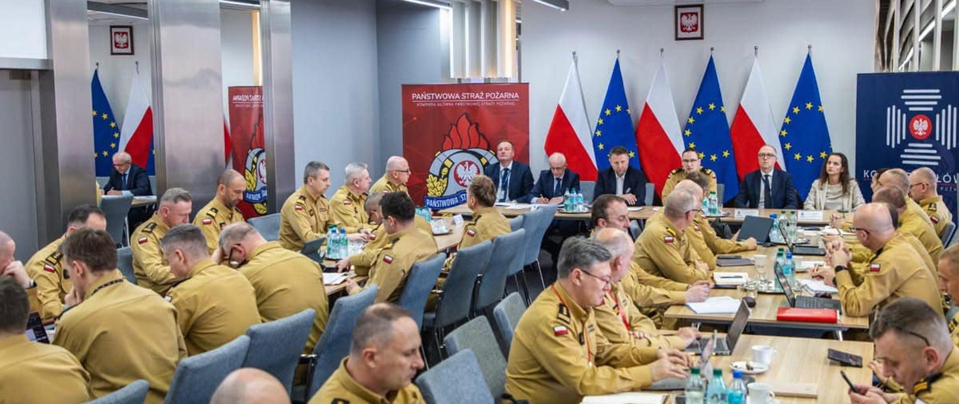Na sali w dwóch rzędach stołów siedzą strażacy w mundurach służbowych. W dali widać stół prezydialny, przy którym siedzą osoby w ubraniach cywilnych. Za nimi w szeregu stoi 6 flag, na przemian biało-czerwona i unijna. Na stołach butelki z wodą, naczynia i dokumenty.