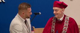Osoba w jasnym garniturze ściska dłoń osoby ubranej w bordowy strój ceremonialny z ozdobnym łańcuchem. W lewej ręce trzyma otwarte etui z medalem. Spotkanie odbywa się w sali z niebieską ścianą i mikrofonem w tle, podczas uroczystości wręczenia odznaczenia.