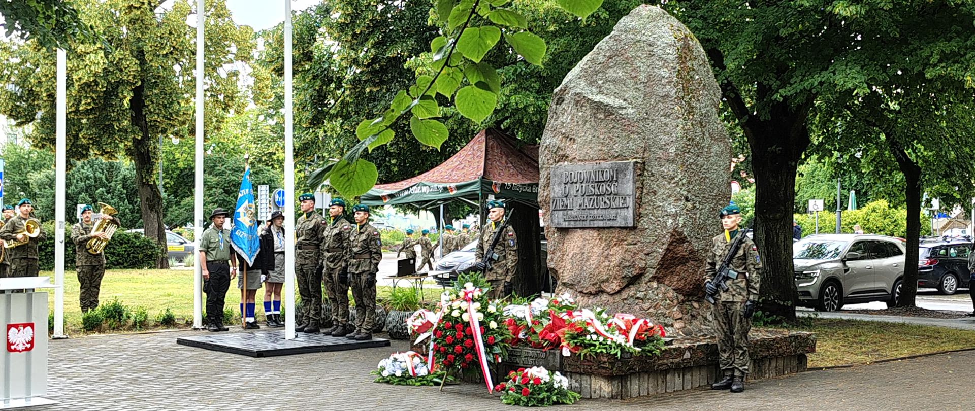 Grupa ludzi w mundurach, warta przy pomniku, przy którym leżą wiązanki biało czerwonych kwiatów z biało-czerwonymi wstęgami.