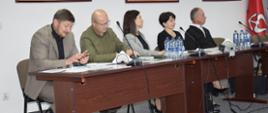 Na zdjęciu widać grupę siedmiu osób podczas spotkania przy długim, drewnianym stole w jasnej sali konferencyjnej. Uczestnicy siedzą po obu stronach stołu, na którym znajdują się dokumenty, butelki i szklanki z wodą, filiżanka kawy oraz telefony.
Na pierwszym planie, po lewej stronie, siedzi mężczyzna w jasnej marynarce Piotr Giza - Kierownik Wojewódzkiego Inspektoratu Ochrony Roślin i Nasiennictwa w Rzeszowie, Oddział w Dębicy, pochylony nad dokumentami. Po jego lewej stronie siedzi kilku mężczyzn w marynarkach. Po przeciwnej stronie stołu znajdują się dwie kobiety oraz mężczyzna w ciemnej marynarce Wójt Gminy Żyraków, który siedzi najbliżej prawej krawędzi zdjęcia.
W tle znajdują się duże okna. Okna są zasłonięte ciemnymi, składanymi panelami. Nad stołem znajdują się prostokątne lampy sufitowe.
