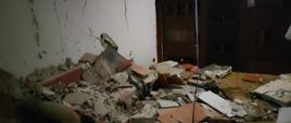 Noc. Widok na zniszczone pomieszczenie. W tle całkowity brak ściany frontowej. Na podłodze pod ścianą w kolorze białym leży sterta gruzu przykrywająca wersalkę. Ściany popękane. Z sufit zwisa żyrandol.