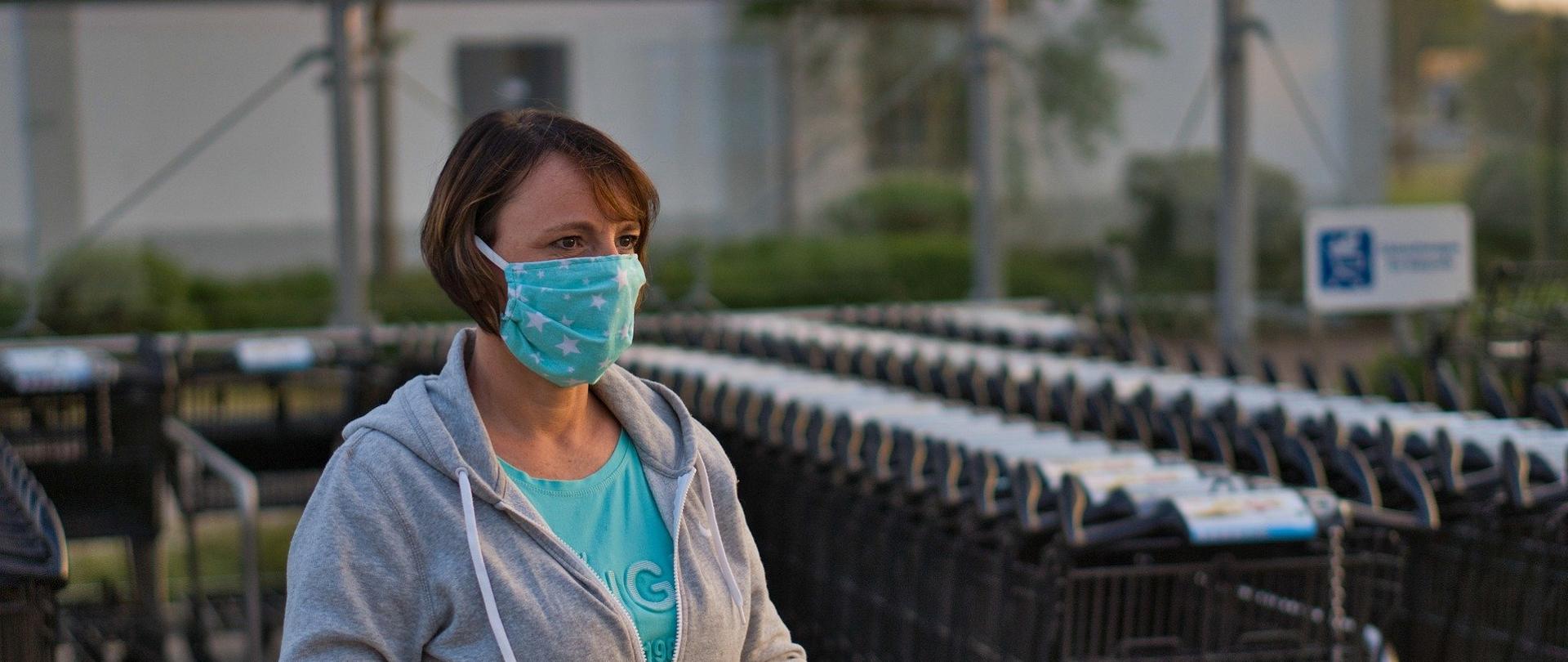 Kobieta w maseczce jednorazowej na twarzy, prowadząca na zewnątrz wózek sklepowy na kółkach
