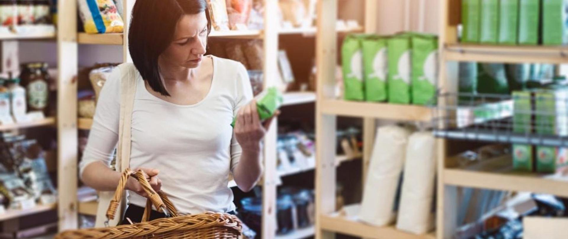 Na zdjęciu widać kobietę z koszykiem wiklinowym w ręce, trzymającą w drugiej ręce produkt w zielonym opakowaniu. Wokół kobiety są drewniane półki sklepowe wypełnione towarami.