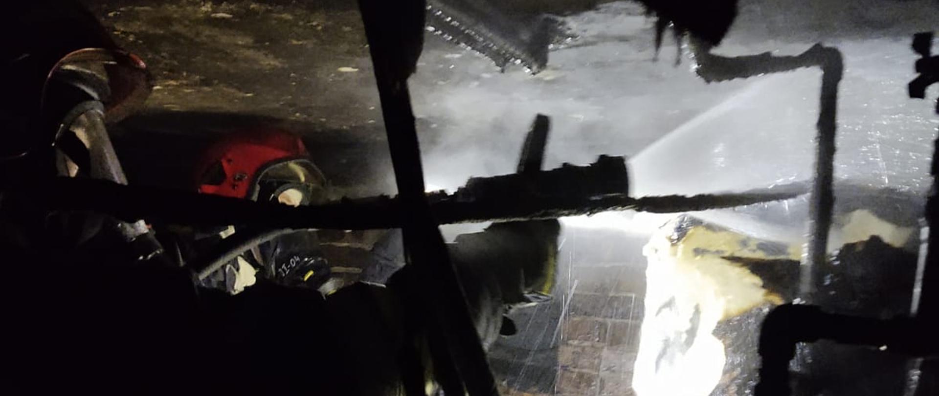 Dwóch strażaków prowadzi działania gaśnicze w piwnicy. Jeden z nich podaje wodę z prądownicy wodnej. Wiadać opalone elementy w piwnicy.