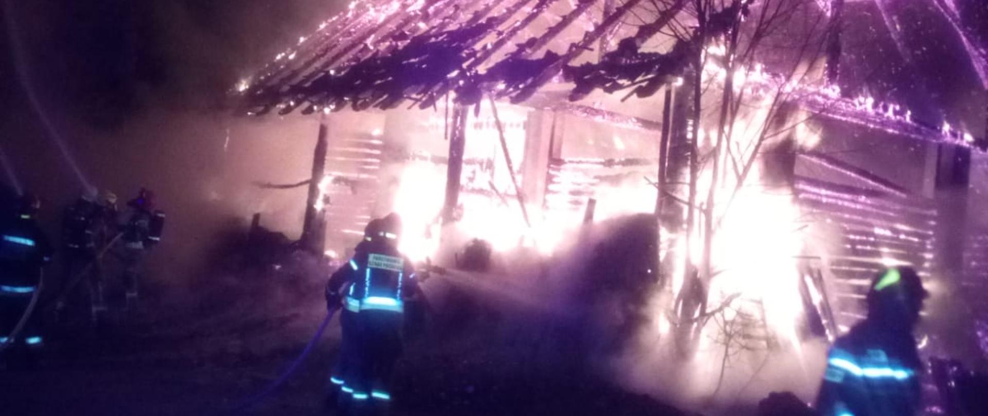 Zdjęcie przedstawia palący się budynek gospodarczy oraz strażaków podających prądy wody.