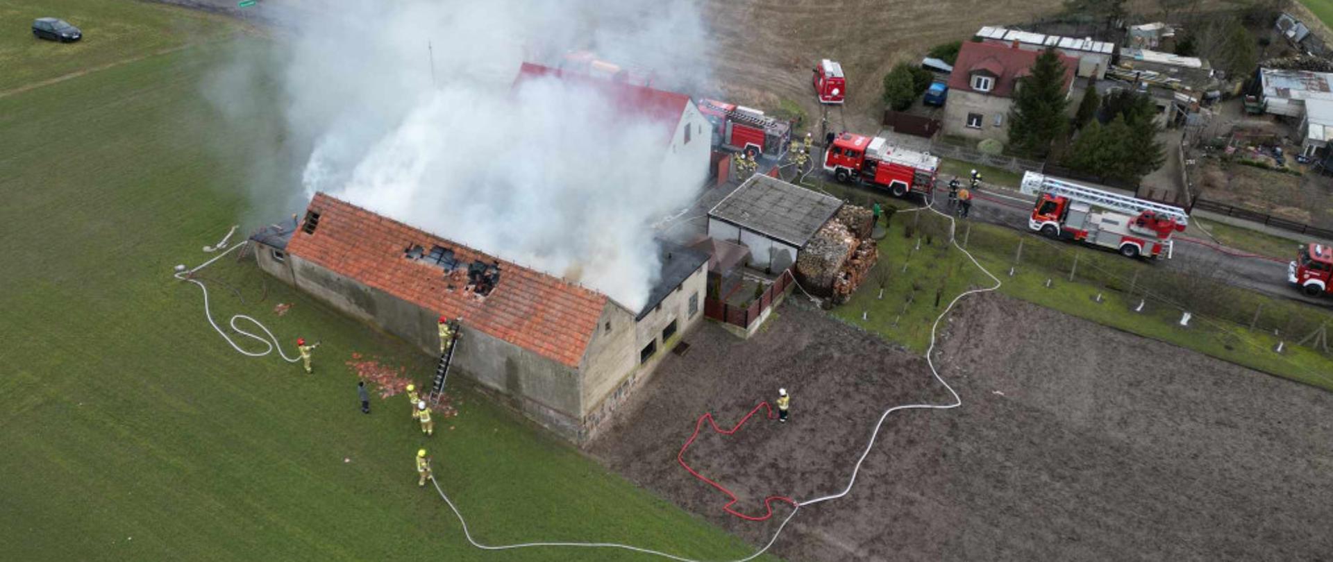 Miejsce działań z lotu ptaka. Z budynku wydobywa się dym. Wozy strażackie stoją na drodze jeden za drugim. Strażacy rozwinęli 3 linie gaśnicze od strony pola. Jeden ze strażaków wchodzi po drabinie 