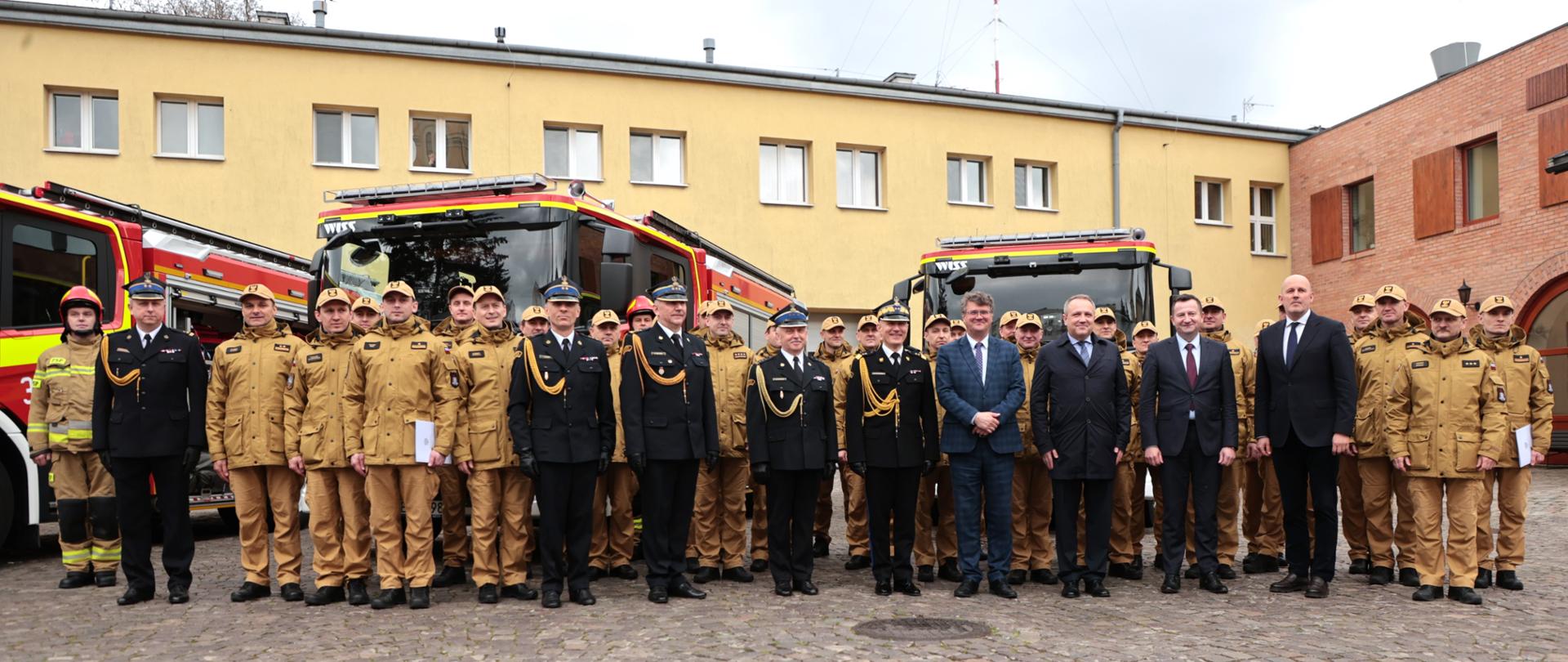 Zbiorowe zdjęcie strażaków i zaproszonych gości , wszyscy stoją w grupie na placu jednostki, w tle samochody rat. - gaś. i budynek strażnicy