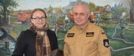 Na tle ściany, na której jest namalowany obraz przedstawiający akcję gaszenia pożaru stoją obok siebie, od lewej: mieszkanka powiatu rawickiego, która wygrała czujnik czadu oraz zastępca komendanta powiatowego PSP. Kobieta ma czarny płaszcz i szalik zawinięty wokół szyi, natomiast strażak - mundur służbowy w kolorze piaskowym. Kobieta trzyma w prawej ręce biały czujnik tlenku węgla. 