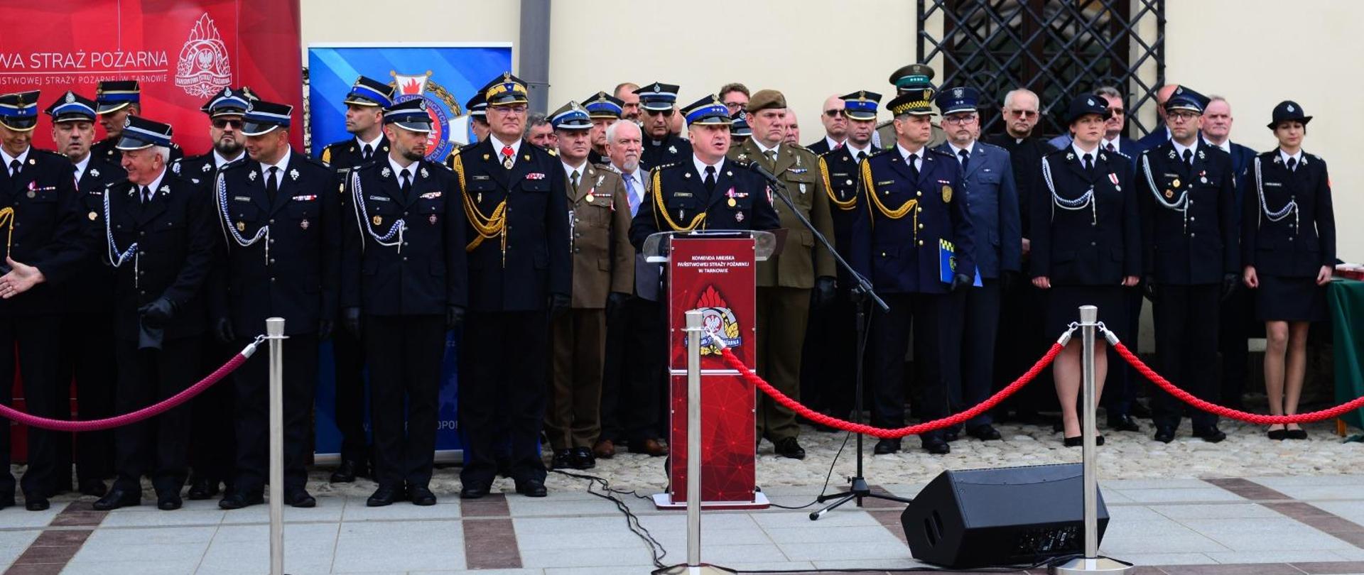 Za mównicą z logiem Państwowej Straży Pożarnej stoją strażacy ubrani w mundury galowe oraz zaproszenie goście z tyłu widać budynek ratusza