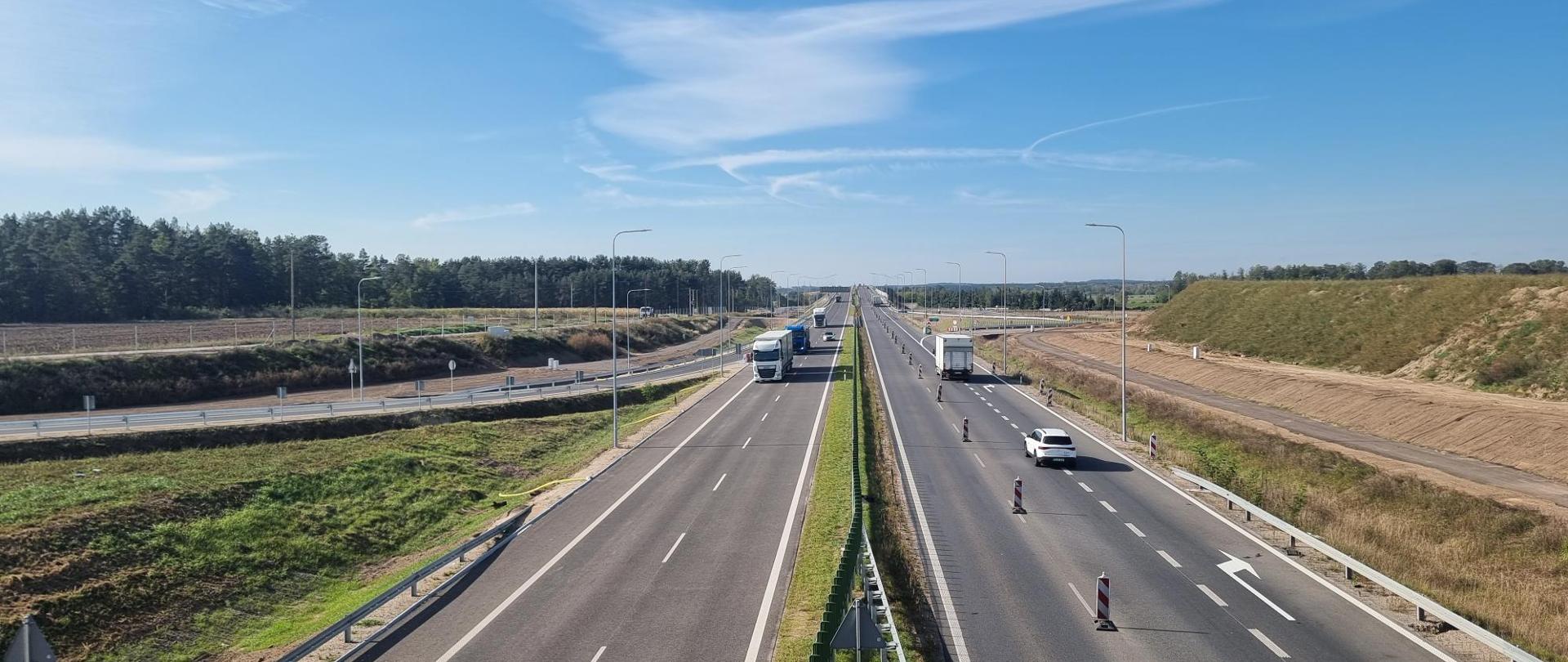 Na zdjęciu widać jezdnię, którą oddziela pas zieleni. Po jezdni jadą samochody osobowe i ciężarowe. Po lewej stronie widać las.