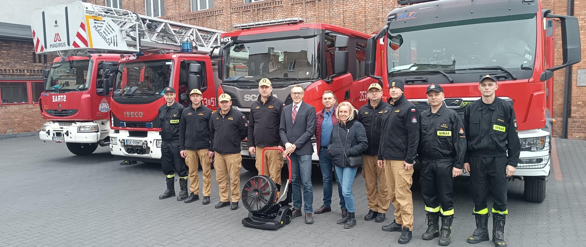 Zdjęcie grupowe Komendanta Miejski Państwowej Straży Pożarnej w Częstochowie wraz z zastępcami i przedstawicielami firmy Brembo Polska oraz strażakami Jednostki Ratowniczo Gaśniczej numer 3 w Częstochowie przed nimi agregat oddymiający.