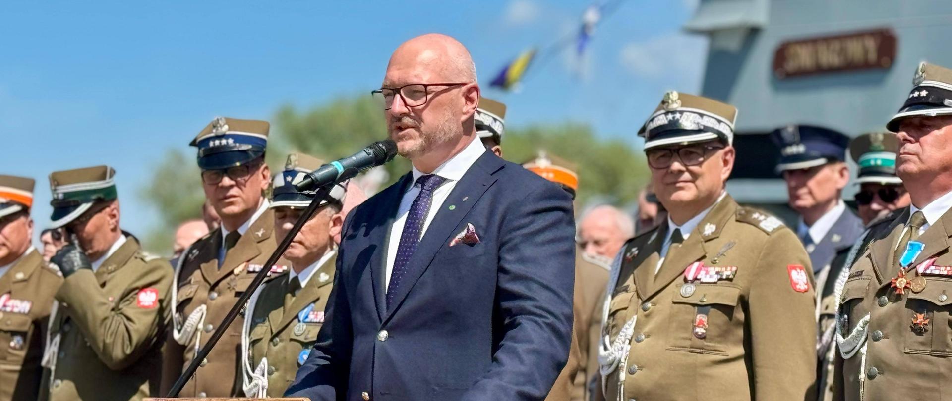 Mężczyzna w garniturze przemawia z mównicy do stojących żołnierzy 