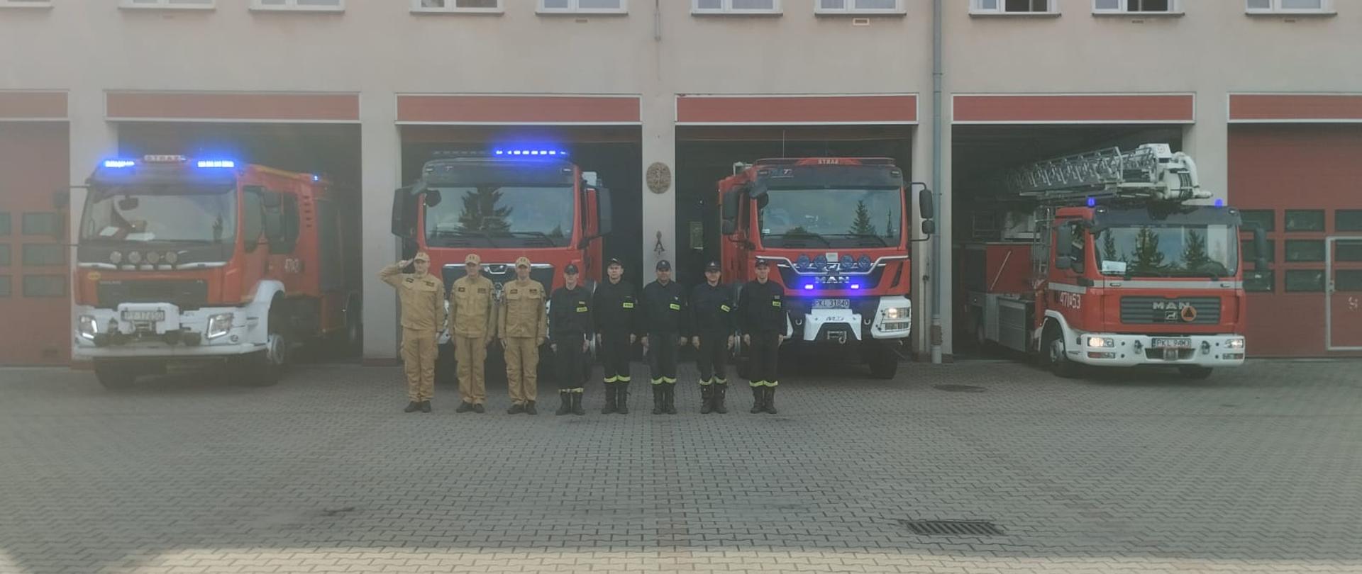Strażacy z JRG Koło oddający hołd Powstańcom Warszawskim. W tle pojazdy pożarnicze z włączoną sygnalizacją świetlną.