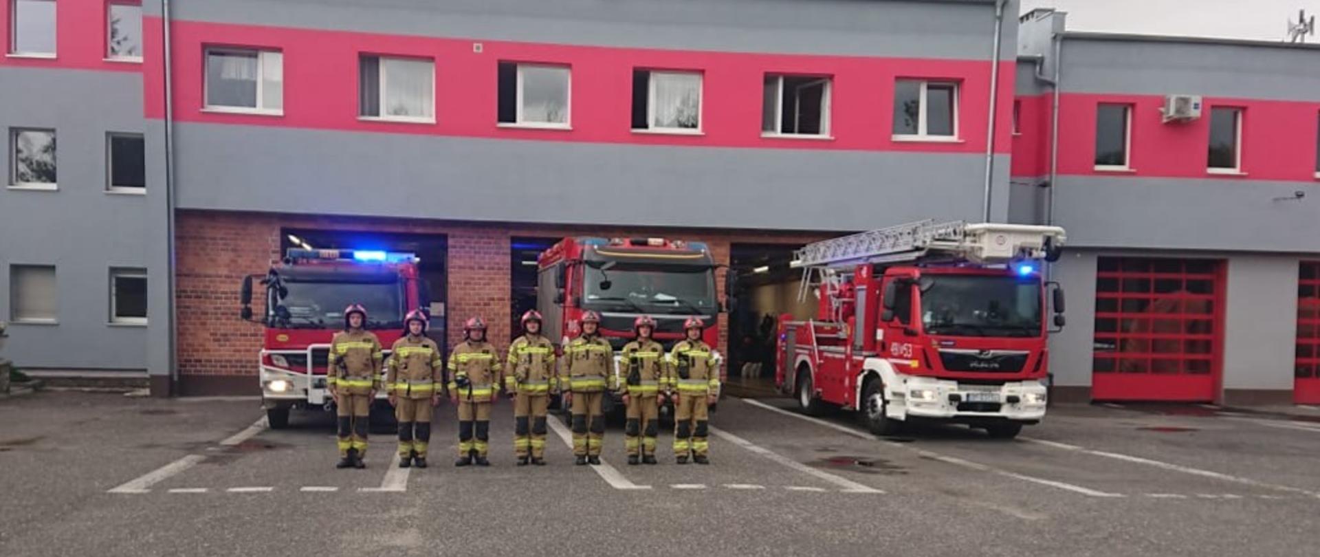 Zdjęcie przedstawia ośmiu strażaków na tle budynku KP PSP w Strzelcach Opolskich i trzech samochodów pożarniczych upamiętniających 77 rocznicę wybuchu Powstania Warszawskiego