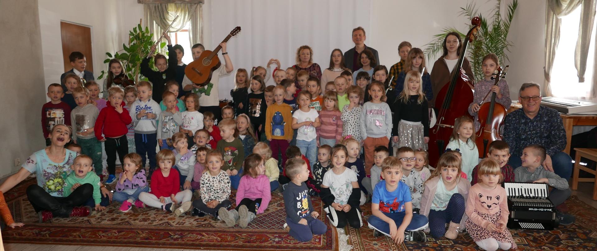 Dzieci z przedszkola, uczniowie ze swoimi instrumentami oraz nauczyciele pozują wspólnie do zdjęcia.