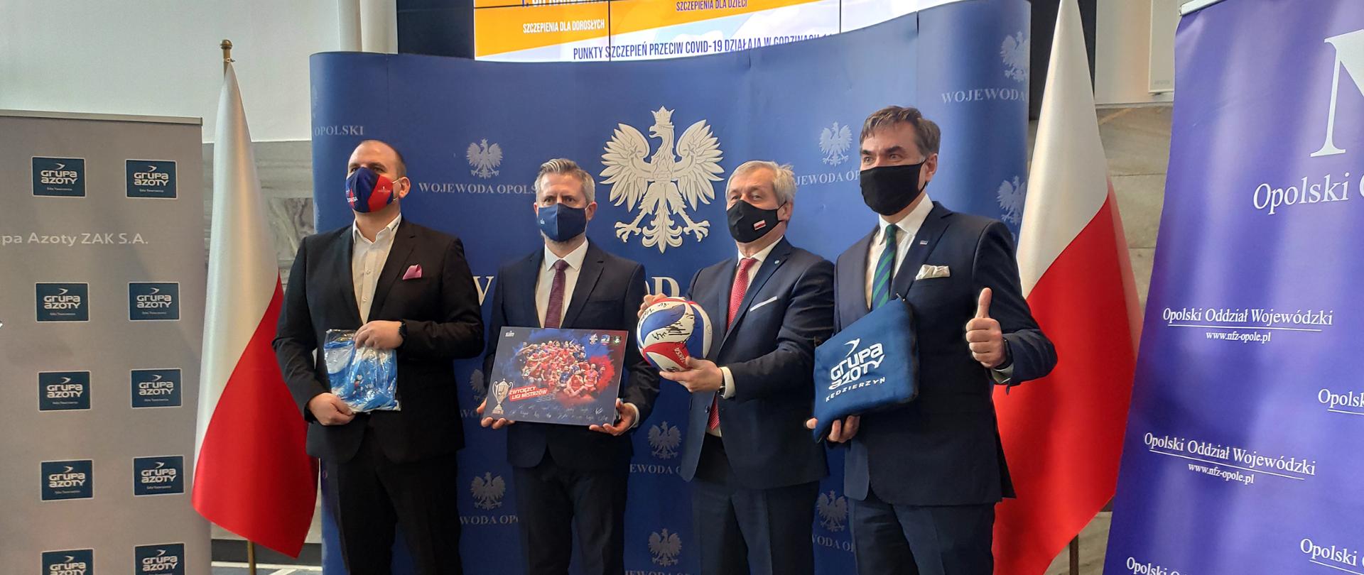 Na zdjęciu wojewoda opolski dyrektor OOW NFZ, prezes Grupy ZAK S.A. oraz ZAKSY. Stoją na tle ścianki granatowej z napisem Wojewoda Opolski, obok biało-czerwone flagi. W tle monitor, na którym wyświetlony jest plakat. 