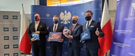 Na zdjęciu wojewoda opolski dyrektor OOW NFZ, prezes Grupy ZAK S.A. oraz ZAKSY. Stoją na tle ścianki granatowej z napisem Wojewoda Opolski, obok biało-czerwone flagi. W tle monitor, na którym wyświetlony jest plakat. 