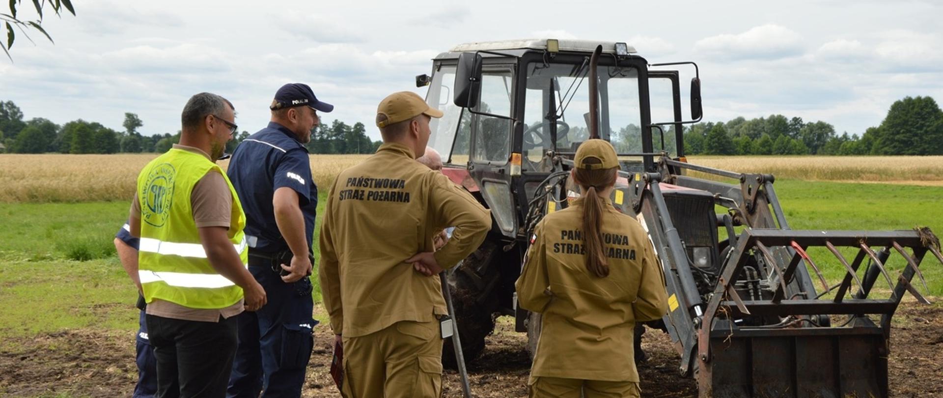 Na zdjęciu widoczna jest grupa pięciu osób stojących na skraju pola uprawnego, obok ciągnika rolniczego wyposażonego w ładowacz czołowy.
Trzy osoby ubrane są w stroje Państwowej Straży Pożarnej w kolorze piaskowym, jedna osoba ma granatowy mundur Policji, a kolejna kamizelkę ostrzegawczą – prawdopodobnie przedstawiciel innej instytucji (np. inspekcji pracy). Wszyscy stoją tyłem lub bokiem do obserwatora i przyglądają się maszynie rolniczej, prowadząc rozmowę lub ocenę jej stanu.
Ciągnik znajduje się na polu, w tle widoczna jest roślinność oraz linia drzew. Pogoda jest umiarkowana, niebo lekko zachmurzone.
Scena przedstawia prawdopodobnie wspólne działania kontrolne lub prewencyjne służb w środowisku rolniczym, związane z bezpieczeństwem pracy i użytkowaniem sprzętu rolniczego.