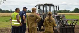 Na zdjęciu widoczna jest grupa pięciu osób stojących na skraju pola uprawnego, obok ciągnika rolniczego wyposażonego w ładowacz czołowy.
Trzy osoby ubrane są w stroje Państwowej Straży Pożarnej w kolorze piaskowym, jedna osoba ma granatowy mundur Policji, a kolejna kamizelkę ostrzegawczą – prawdopodobnie przedstawiciel innej instytucji (np. inspekcji pracy). Wszyscy stoją tyłem lub bokiem do obserwatora i przyglądają się maszynie rolniczej, prowadząc rozmowę lub ocenę jej stanu.
Ciągnik znajduje się na polu, w tle widoczna jest roślinność oraz linia drzew. Pogoda jest umiarkowana, niebo lekko zachmurzone.
Scena przedstawia prawdopodobnie wspólne działania kontrolne lub prewencyjne służb w środowisku rolniczym, związane z bezpieczeństwem pracy i użytkowaniem sprzętu rolniczego.