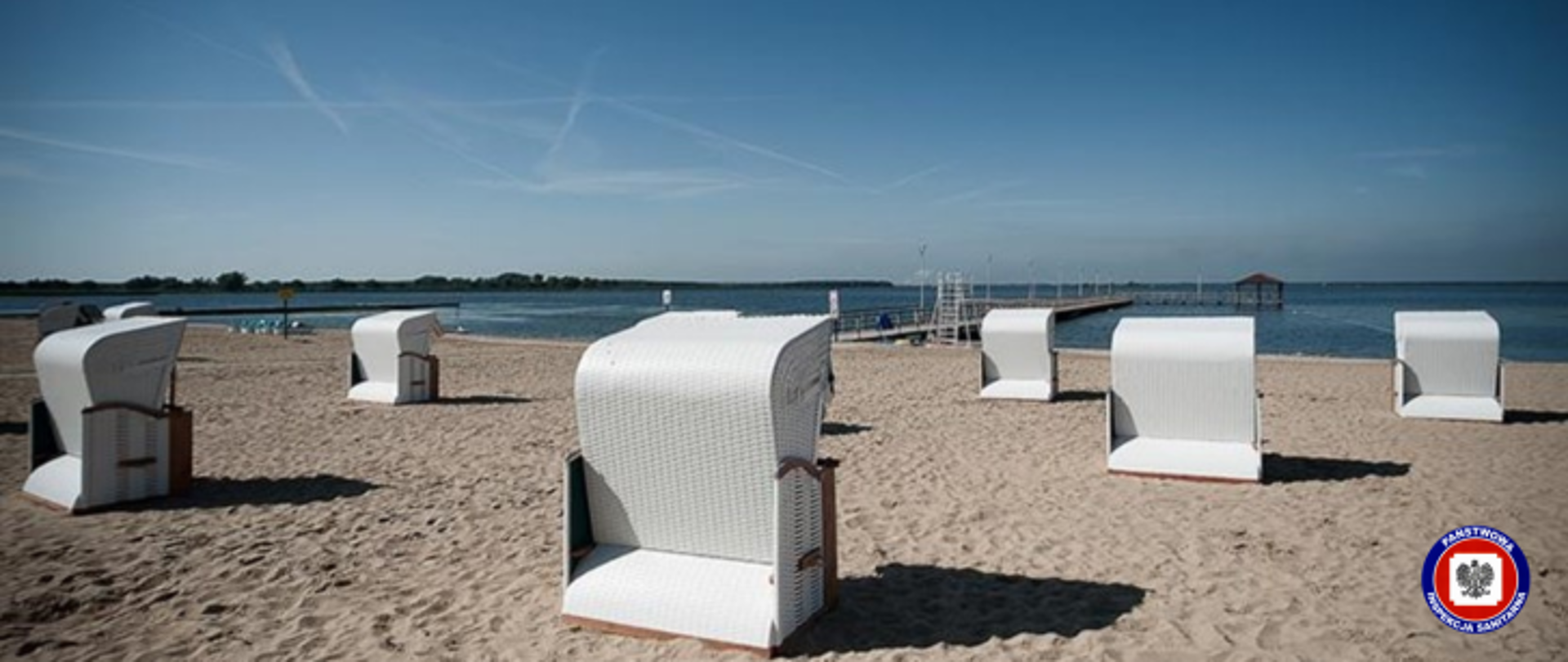 Na plaży, na piasku, stoi tyłem osiem wiklinowych koszy plażowych przeznaczonych do odpoczynku w kolorze białym . w tle akwen wodny z molo. Niebo jest bezchmurne i niebieskie. W lewym dolnym rogu logo Inspekcji Sanitarnej.