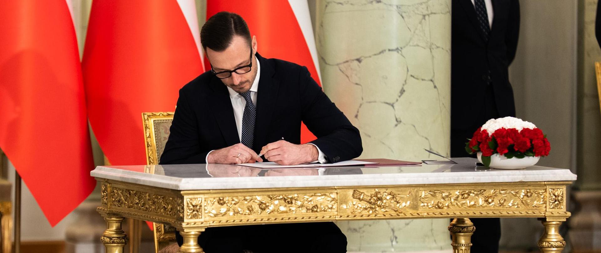 Minister Jakub Jaworowski siedzi przy stole i podpisuje dokument. Na stole stoją kwiaty. W tle biało-czerwone flagi.