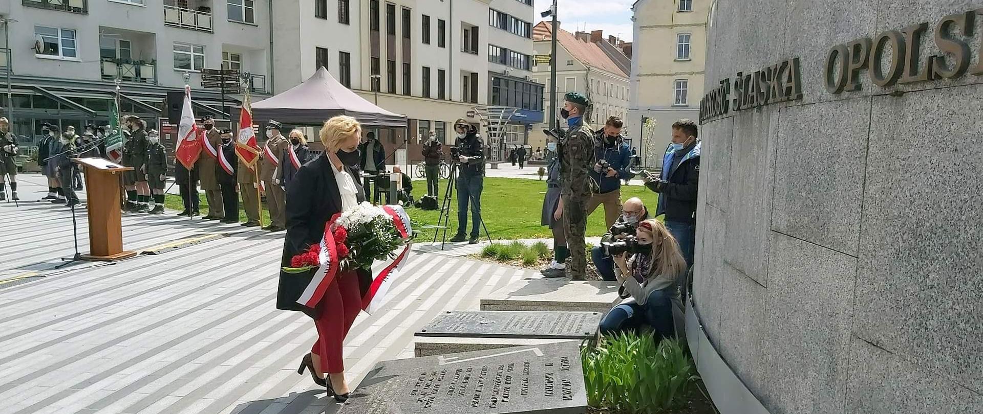 Wicewojewoda opolski podczas uroczystości, upamiętniających 76. rocznicę zakończenia II Wojny Światowej. Na zdjęciu wicewojewoda (po prawej stronie) idąca z floretem w kierunku pomnika na Placu Wolności w Opolu. Przed nią, na ziemi, 3 tablice upamiętniające wydarzenia sprzed lat. Na przeciw wicewojewody, po pomnikiem, fotoreporterzy. W tle pozostali uczestnicy spotkania oraz budynki, zalkalizowane przy ul. Krakowskiej w Opolu. 
