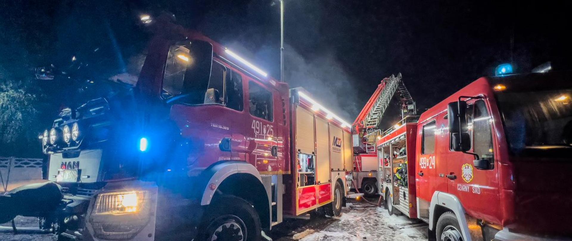 Zdjęcie przedstawia wozy strażackie stojące na terenie akcji. Samochody mają włączone światła błyskowe, i oświetlają teren prowadzonych działań.