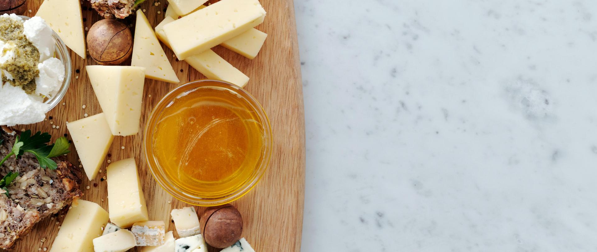 Zdjęcie przedstawia klasyczną Deskę serów. Na drewnianej desce widzimy pokrojony ser żółty, pleśniowy, biały typu greckiego. Szklankę z miodem. orzechy oraz kromkę chleba wieloziarnistego.