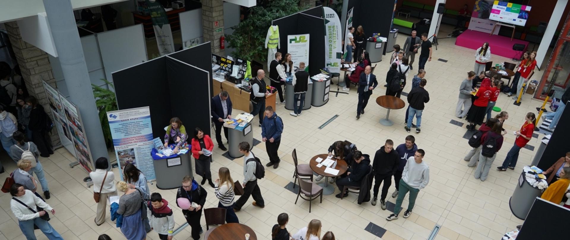 Duży hol w budynku publicznym, prawdopodobnie na uczelni, wypełniony młodzieżą i dorosłymi podczas festiwalu umiejętności. Wiele stoisk informacyjnych oddzielonych czarnymi parawanami jest rozmieszczonych na jasnej posadzce, a uczestnicy rozmawiają i oglądają broszury.