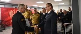 Strażak w mundurze Państwowej Straży Pożarnej ściska dłoń mężczyzny w garniturze podczas uroczystości w sali konferencyjnej. W tle stoją inni strażacy w mundurach galowych i służbowych, a na ścianie widoczny jest czerwony baner z godłem Polski i napisem „Państwowa Straż Pożarna”.