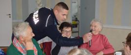 Spotkanie z seniorami. Sala w jednej z rawickich restauracji. Pośród kilku uczestniczek spotkania stoi policjant, który prezentuje seniorkom legitymację służbową. Pani siedząca w środku ma okulary.