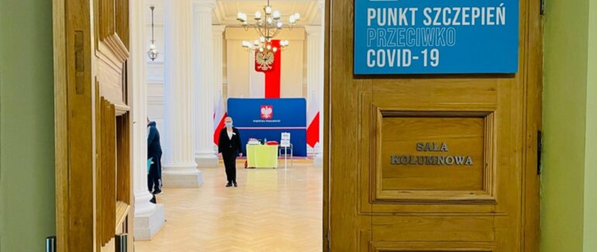 Wejście do sali kolumnowej Podkarpackiego Urzędu Wojewódzkiego w Rzeszowie. Na drzwiach niebieska tablica z napisem: Punkt szczepień przeciwko COVID-19. W tle wnętrze sali kolumnowej i kobieta w maseczce
