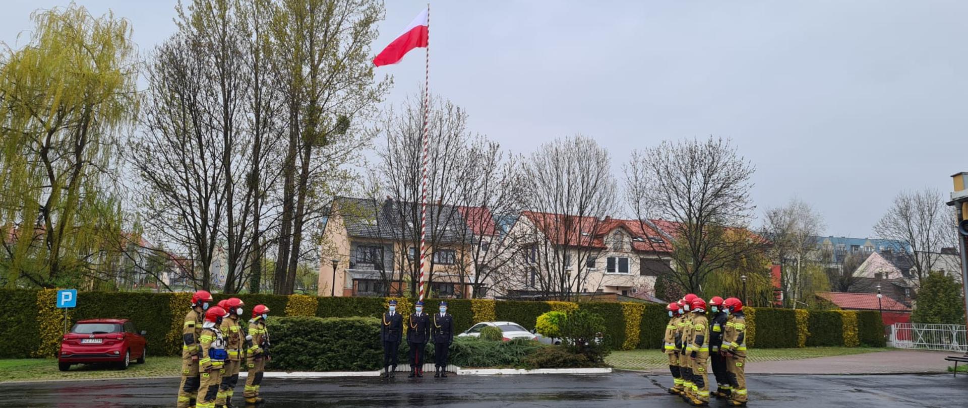 Na zdjęciu strażacy stoją na placu strażnicy w dwuszeregu ubrani w ubrania specjalne koloru piaskowego. Jedna zmiana z prawej druga z lewej strony zdjęcia. Po środku maszt na którym znajduje się flaga Polski z przed masztem poczet flagowy ubrani w ubrania wyjściowe.