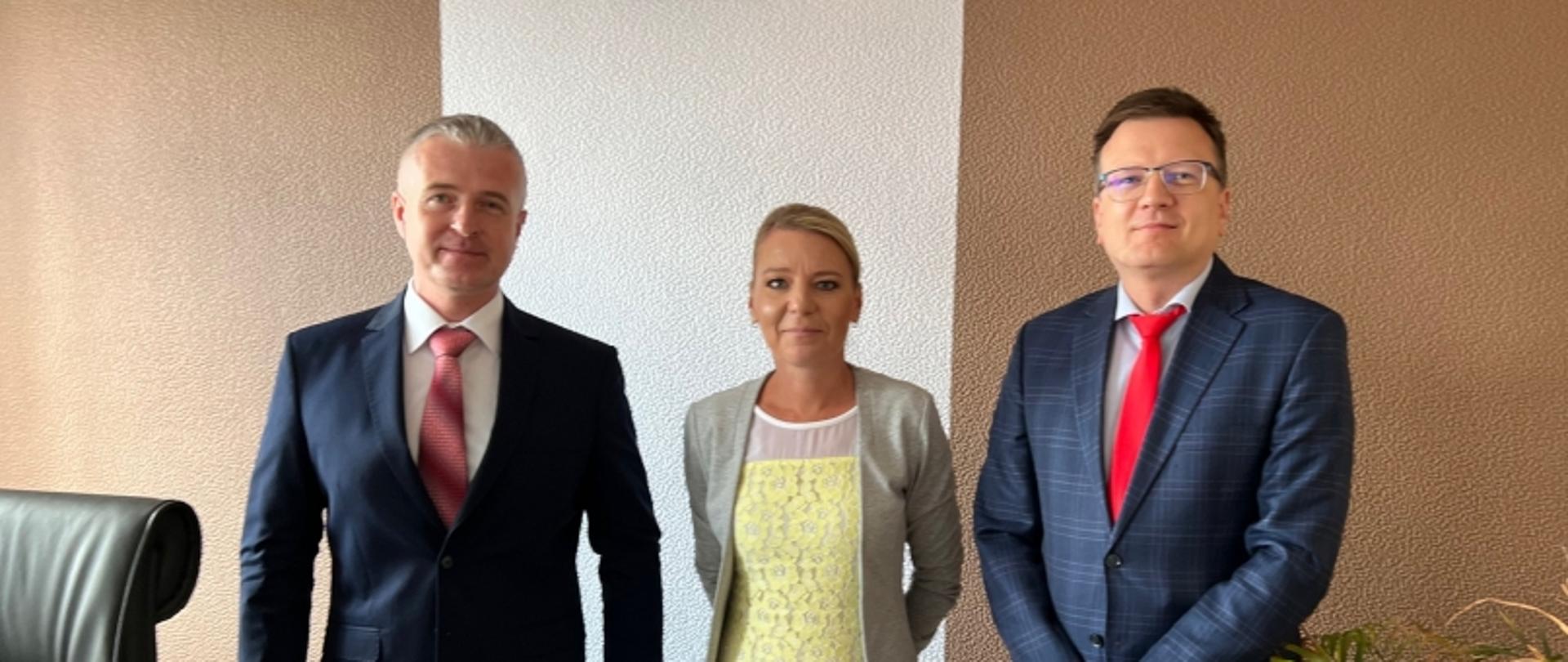 Trzy osoby stojące obok siebie; dwie w garniturach z czerwonymi krawatami, jedna w jasnym stroju, w tle fakturowana ściana.