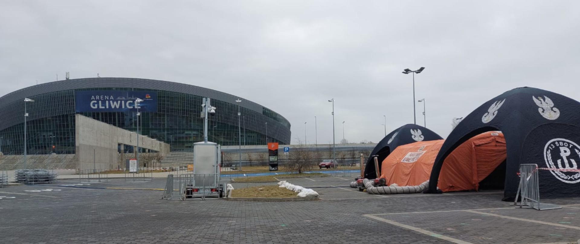 Na zdjęciu widoczne na parkingu przed halą widowiskową, po prawej stronie trzy namioty. W środku pomarańczowy ,pneumatyczny PSP, po bokach namioty WOT. Na wprost widoczna hala Arena w Gliwicach.