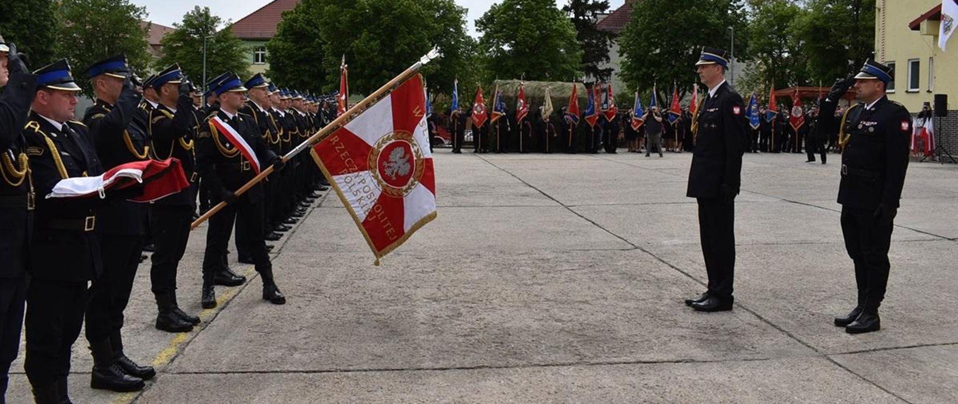 Dzień. Widok na plac manewrowy. Z lewej strony stoi kompania reprezentacyjna składająca się z funkcjonariuszy PSP. Obok kompani stoi poczet flagowy oraz poczet sztandarowy, który prezentuje sztandar. Przed sztandarem stoi dwóch funkcjonariuszy PSP. Wszyscy strażacy ubrani są w mundury wyjściowe ze złotym sznurem galowym oraz rogatywki. W głębi zdjęcia w szeregu stoi kilkanaści innych pocztów sztandarowych. 