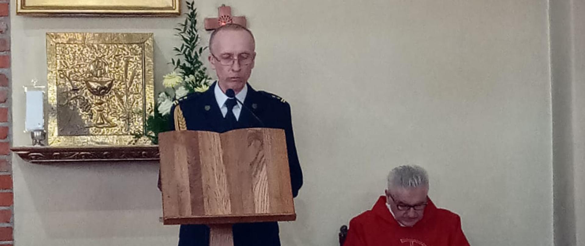 Zdjęcie przedstawia mężczyznę podczas czytania na mszy świętej. Obok niego znajduję się Ksiądz.