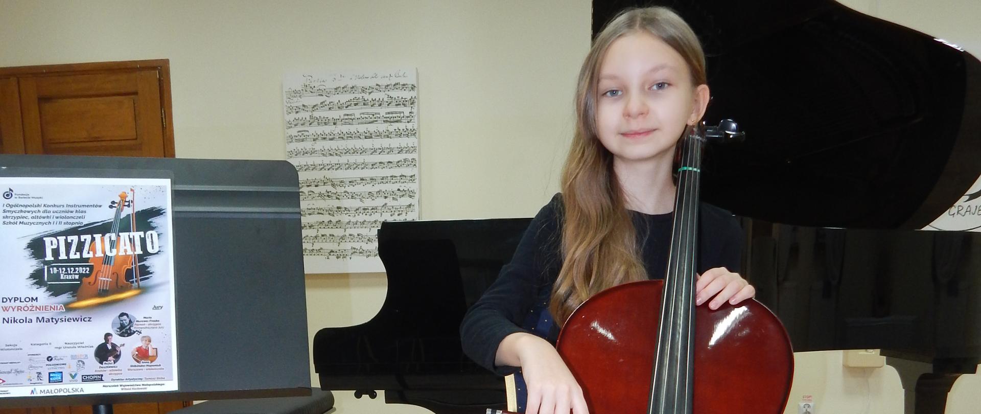 Dziewczynka ubrana w czarną bluzkę i szarą spódnicę siedzi przy wiolonczeli, obok stoi pulpit z dyplomem za zdobycie Wyróżnienia kategorii II w I Ogólnopolskim Konkursie Instrumentów Smyczkowych dla uczniów klas skrzypiec, altówki i wiolonczeli Szkół Muzycznych I i II stopnia "PIZZICATO" w Krakowie (edycja on-line). W tle fortepian i obrazy.