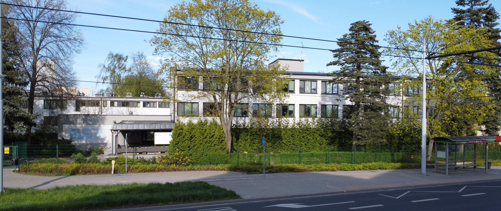 Na zdjęciu widać budynek Bursy widziany do frontu. Budynek jest jasnoszary, z prawej strony widać dwa piętra z ciemnymi oknami, z lewej strony widać wejście przykryte szarym dachem i na ścianie niebieskie logo BSA. Z tyłu widać drugi podobny budynek. Z przodu widać fragment pasa jezdni z białymi oznaczeniami, po prawej stronie przystanek autobusowy. Przed budynkiem i wokół niego dużo zielonych drzew i krzewów.