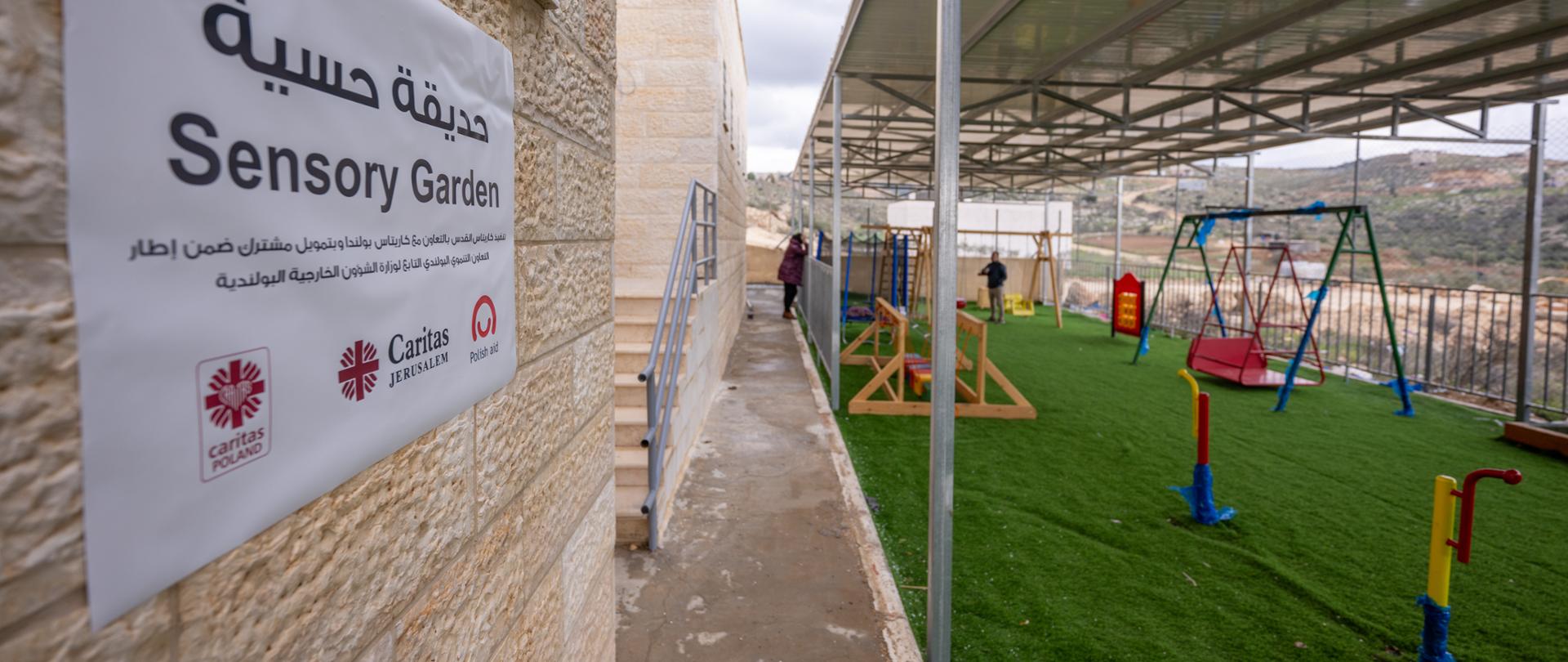 Na dalszym planie dwoje dorosłych osób stoi na zadaszonym i ogrodzonym placu zabaw dla dzieci z niepełnosprawnościami