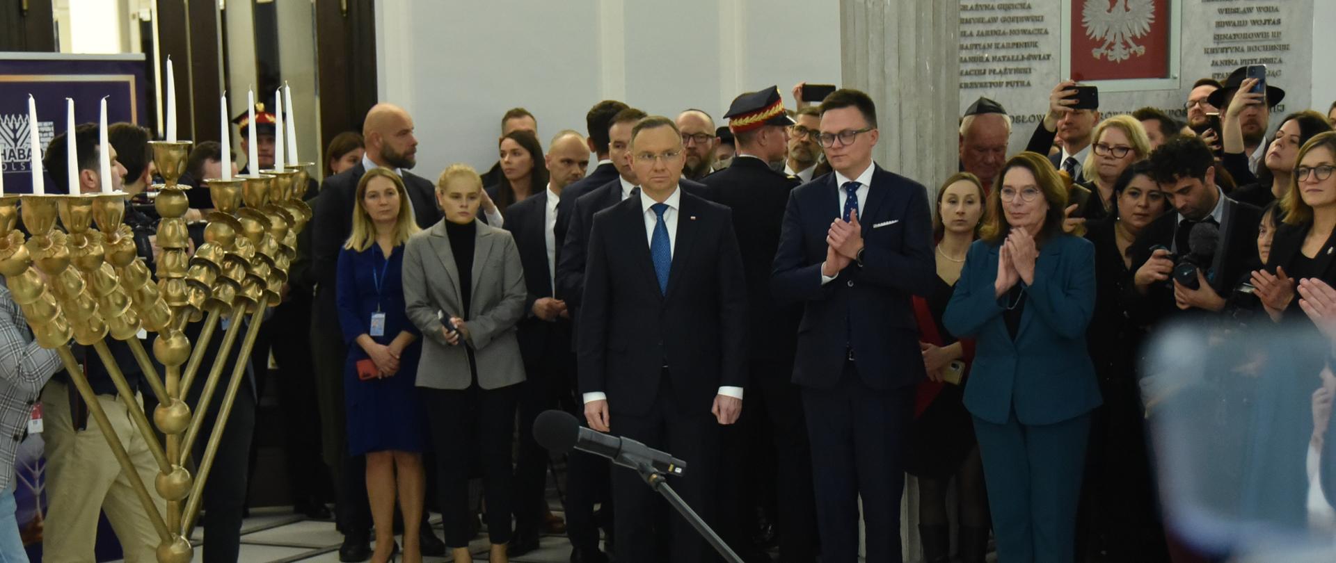 W białej sali stoi dużo ludzi, pośrodku marszałek Hołownia i prezydent Duda, przed nimi złoty dziewięcioramienny świecznik.