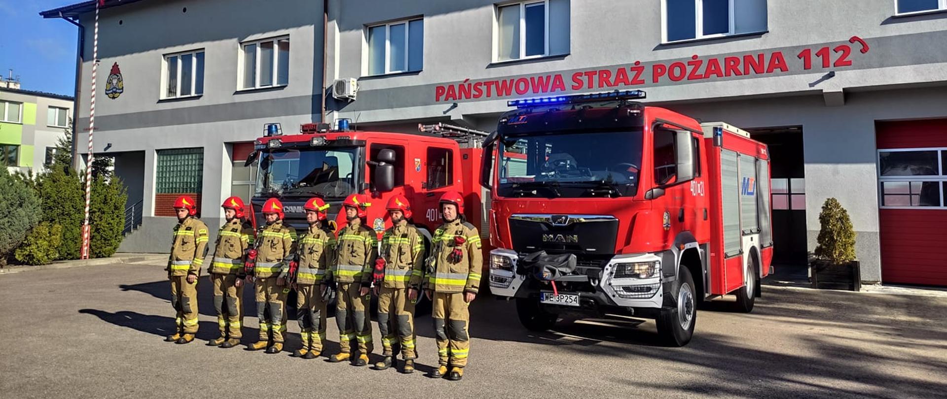 strażacy stoją przed samochodami pożarniczymi, które włączone mają sygnalizacje świetlne i dźwiękowe
